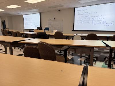 photo of classroom taken from the back of the class showing tables, chairs, two screens on front wall